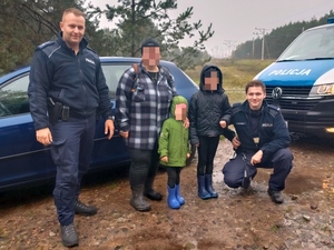 Policjant w mundurze, kobieta w kraciastej kurtce, dziecko w zielonej kurtce, dziecko w granatowej kurtce i kucający policjant w mundurze znajdujący się na tle oznakowanego radiowozu i granatowego samochodu