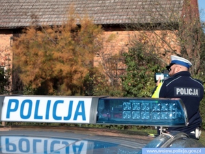 na pierwszym planie belka od świateł błyskowych w radiowozie z napisem policja, w tle  policjant ruchu drogowego wykonujący pomiar prędkości