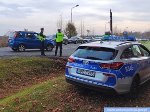 Zdjęcie przedstawia na pierwszym planie po lewej stronie radiowóz oznakowany stojący tyłem, w stoją przy drodze dwaj policjanci ruchu drogowego w kamizelkach odblaskowych z napisem Policja oraz w białych czapkach, za nimi stoją samochody