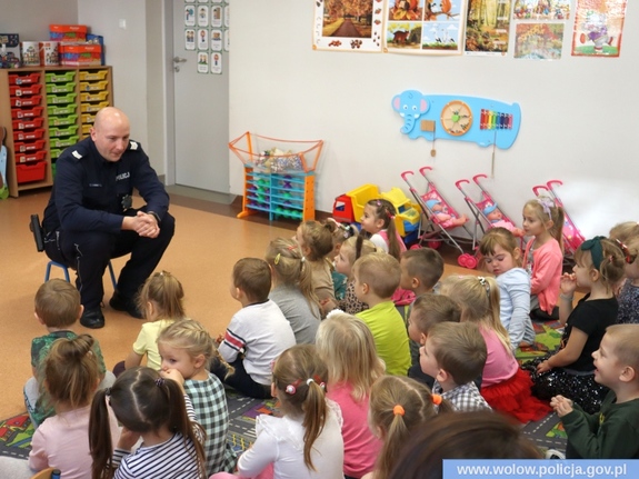 Zdjęcie przedstawia policjanta siedzącego w pomieszczeniu na małym krzesełku przed grupą dzieci w wieku przedszkolnym