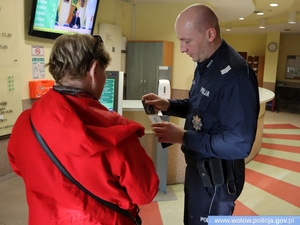 w pomieszczeniu policjant w mundurze prezentuje kobiecie w czerwonej kurtce nakładkę zabezpieczającą na kartę płatniczą