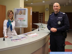 W pomieszczeniu policjant w mundurze stoi przy kontuarze, za kontuarem stoi kobieta z apaszką i białą bluzką, z tyłu wisi plakat ze starszą kobietą i napisem nie daj się oszukać