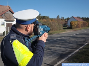 policjant w mundurze i białej czapce trzyma w ręku miernik prędkości, w tle zabudowania