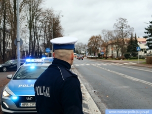 policjant w mundurze i białej czapce stoi przy drodze, w tle oznakowany radiowóz