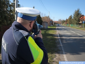 policjant w białej czapce stojący przy drodze trzymający miernik prędkości