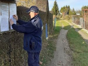 Na zdjęciu widzimy umundurowanego policjanta, który przyczepia ogłoszenie na terenie ogródków działkowych.