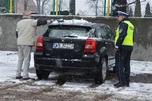 Na zdjęciu widzimy mężczyznę, który odśnieża samochód koloru czarnego, a obok stoi policjant ruchu drogowego w kurtce, czapce oraz kamizelce odblaskowej.